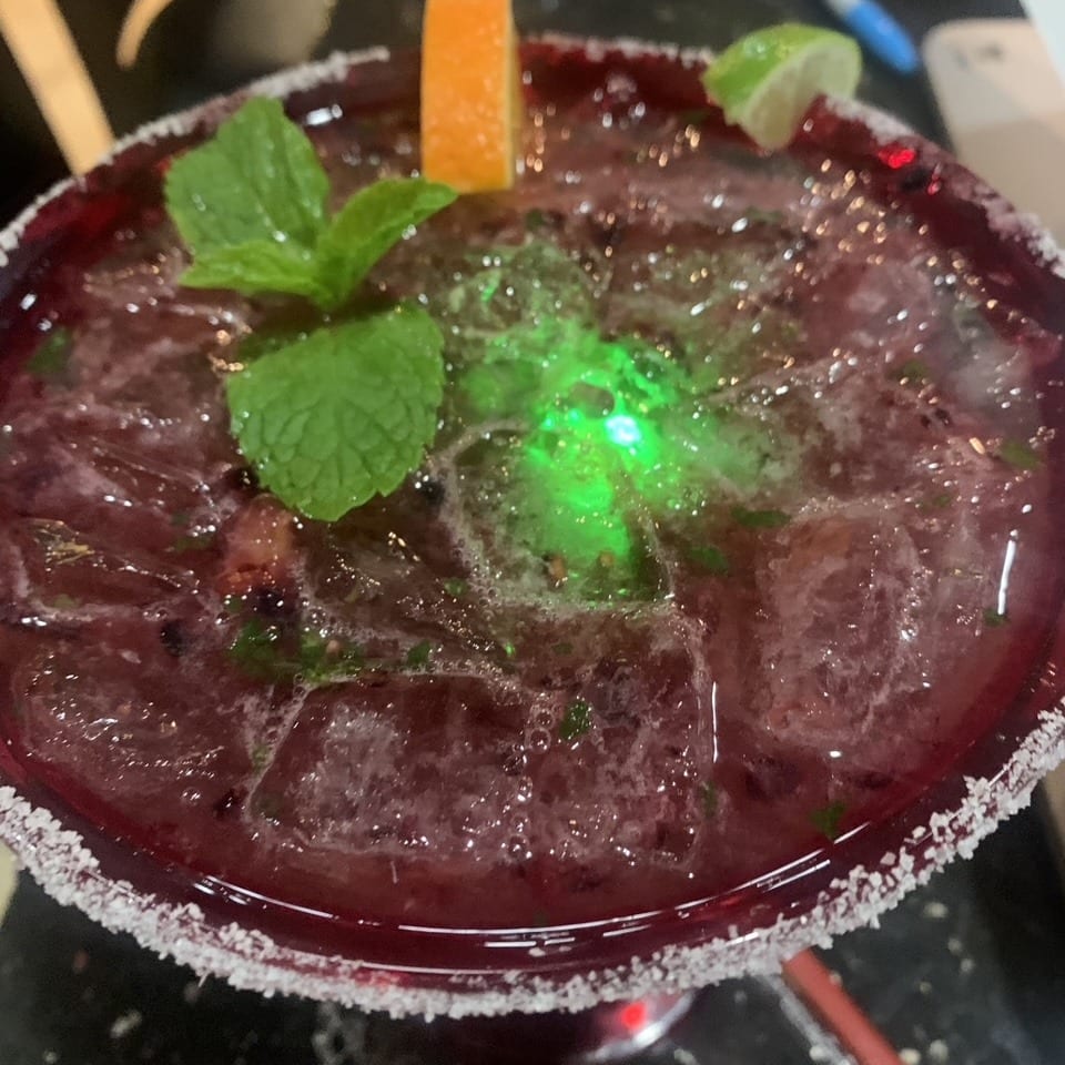 Red iced cocktail in a salt-rimmed glass, garnished with mint, orange wedge, and lime.