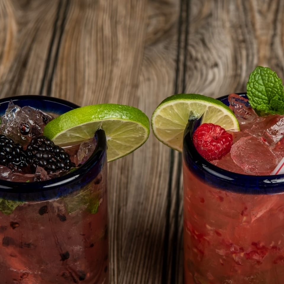 Two berry mojitos with lime slices, mint leaves, ice, and striped straws on a rustic wooden table.