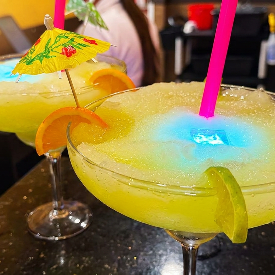 Two bright yellow cocktails garnished with citrus wedges, one topped with a paper umbrella and blue glow, sit on a bar counter.